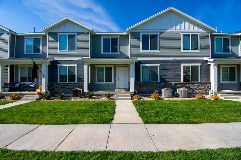 A row of modern, two-story townhouses under a blue sky. Each unit features gray siding, white trim, and a small front porch. A concrete walkway leads through a well-manicured green lawn adorned with neatly arranged plants and shrubs.