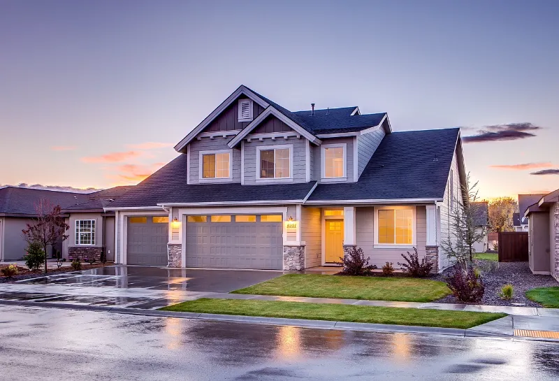 Suburban house at dusk with a lit interior, featuring a double garage, peaked roof, and a neatly manicured lawn. The driveway and street are wet, reflecting the soft glow of lights. The sky is a mix of blue and orange hues.