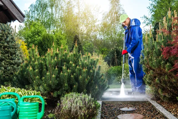 A person wearing a blue raincoat and a green cap uses a pressure washer on a garden pathway surrounded by lush greenery. Two green watering cans sit in the foreground. Sunlight filters through the trees in the background.