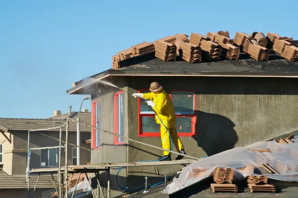 A construction worker in a yellow suit power washes the exterior of a house. The roof is covered with stacks of brown tiles. The house has red-framed windows, and other houses are visible in the background under a clear blue sky.