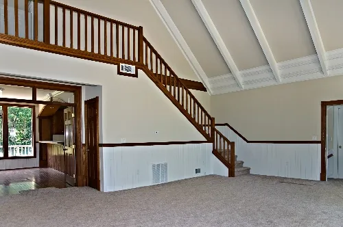 Spacious room with vaulted ceiling and wood accents. Stairs with wooden railing lead to an upper level. Carpeted floor and an open doorway reveal a glimpse of another room with hardwood floors and large windows. Walls feature white paneling and trim.