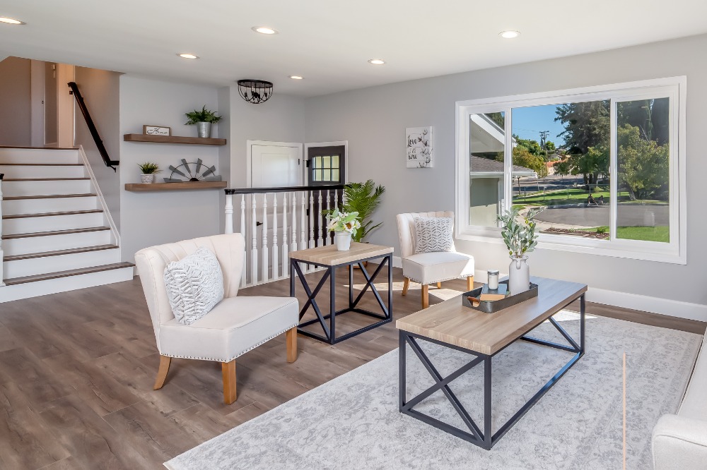 Modern living room with large window, showcasing a view of a suburban street. Two beige armchairs and two wooden coffee tables with black metal frames sit on a light rug. White staircase to the left, and decorative shelves on the wall.