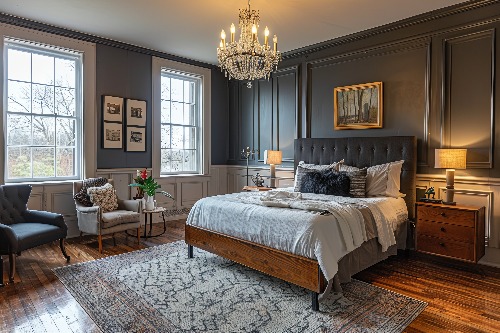 Elegant bedroom with a dark accent wall, featuring a chandelier, large windows, and a tufted bed. A cozy seating area on the left includes a plush chair. Warm lighting from lamps complements the classic decor.