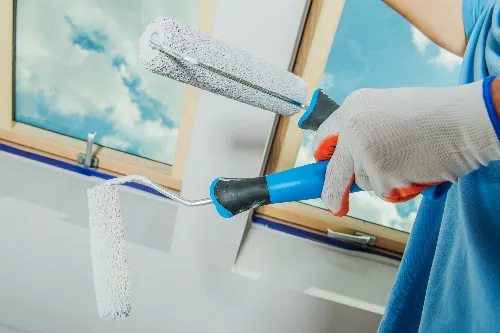 A person wearing gloves holds two paint rollers, one large and one small, near a partially painted wall and a window with a view of the blue sky and clouds. The paint appears to be white.