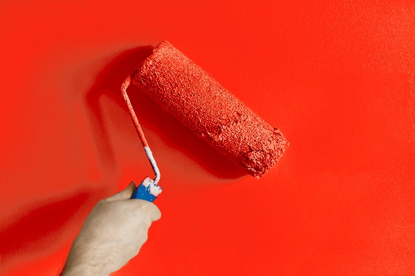 A hand holding a paint roller applies bright red paint to a wall. The roller is covered in red paint, and the entire image is dominated by the vivid red color of the wall.