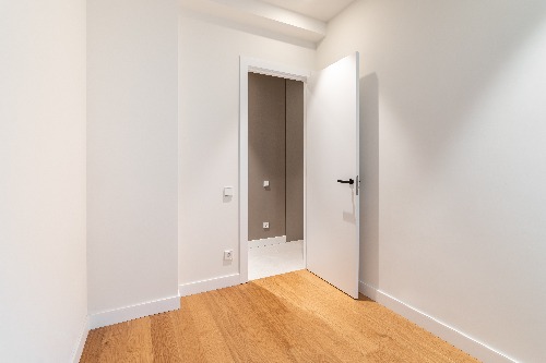 A small, empty room with white walls and a wooden floor. An open door leads to another room with beige walls and a tiled floor. Natural light enters from outside the frame, casting soft shadows on the floor.