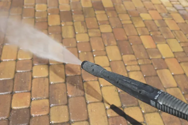 A pressure washer is being used to clean a brick pavement. Water is spraying forcefully from the nozzle, creating a misty effect against the surface of the bricks beneath.