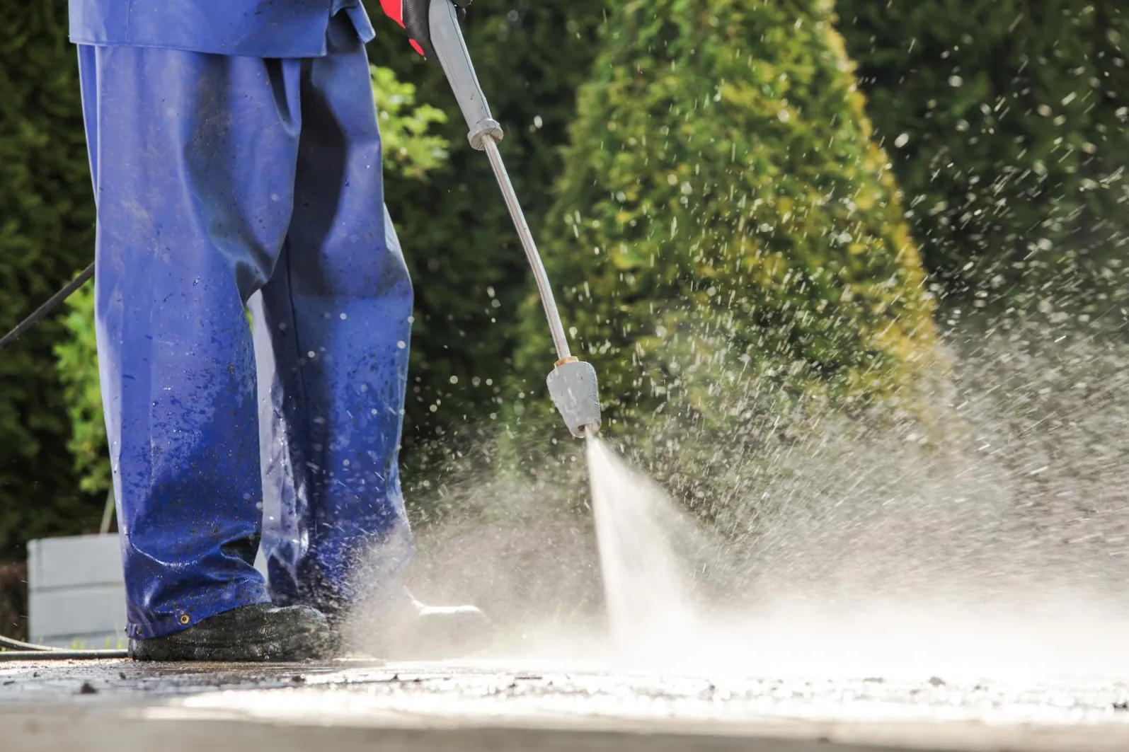 A person in blue protective clothing uses a pressure washer to clean a surface. Water sprays forcefully from the nozzle, creating mist and droplets. Green foliage is blurred in the background.